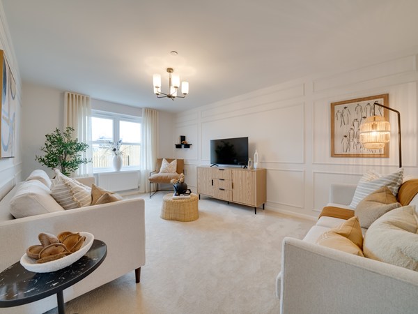 A bright, spacious living room decorated in neutral tones with soft beige carpets and cream walls featuring elegant panel detailing. Two upholstered sofas with assorted cushions face each other across a central woven circular coffee table. A wooden sideboard with a television sits against the far wall, with framed abstract artwork hanging above and to the right. A large window with light curtains lets in natural light, decorative elements, and a small side table complete the modern, calming design.