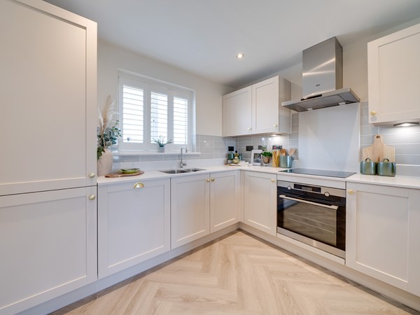 Corner shaped kitchen with light grey cupboards. There is an oven, hob and cooker hood and a sink with a window above