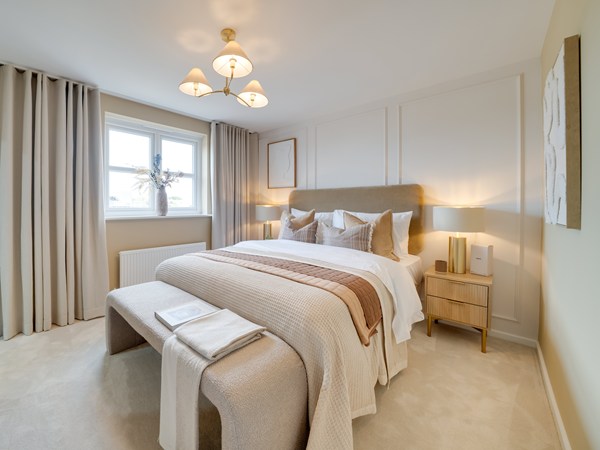A bright, neutral-toned bedroom featuring a large bed with layered bedding in cream, beige, and taupe. The bed has multiple decorative cushions and a soft upholstered headboard. A textured bench sits at the foot of the bed with folded towels placed on top. Matching bedside tables with lamps flank the bed, and long floor‑to‑ceiling curtains frame a window that lets in natural light. The walls are decorated with subtle panel detailing and a simple framed artwork, creating a calm and elegant atmosphere.