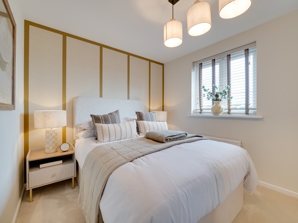 A modern bedroom with a neatly made bed featuring white bedding, a quilted throw, and striped cushions. A tall upholstered headboard stands against a wall with gold panel detailing. A bedside table with a lamp and small decor sits on the left. On the right, a window with wooden blinds and a vase of greenery lets in natural light. Three pendant lights hang from the ceiling, and the room is softly carpeted.