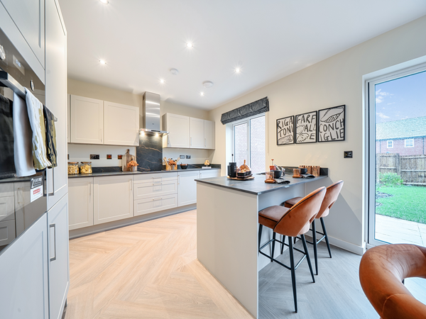 Modern kitchen with white cabinets, grey worktops, integrated appliances, a central cooking area with stainless steel hood, and a breakfast bar with orange bar stools overlooking the garden through a glass door.