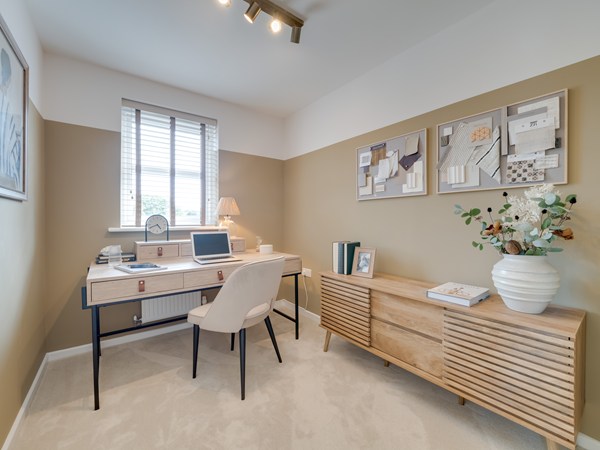 A modern home office with a white desk and a laptop, books, and a small lamp arranged neatly on top. A cushioned chair sits at the desk beneath a window with wooden blinds. On the right, a wooden sideboard holds a large vase with greenery and decor. Above it, a pinboard displays fabric samples and notes. The room has soft carpeting, two‑tone walls, and ceiling spotlights providing warm light.