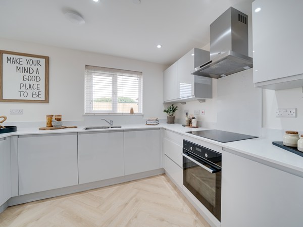 A modern white kitchen with glossy cabinets, a built‑in oven, an electric stovetop, and a stainless steel range hood. A sink sits beneath a window with white blinds, and light wood herringbone flooring runs throughout. A framed sign on the wall reads “MAKE YOUR MIND A GOOD PLACE TO BE,” and small décor items like a plant and cutting board add warmth.