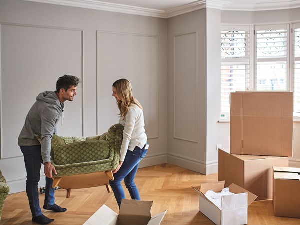 Couple carry a green armchair sofa together in a new build living room with parquet floors, moving boxes and grey walls