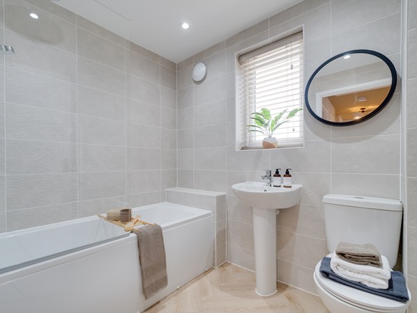 A modern bathroom with light grey tiled walls, a white bathtub on the left with a wooden bath tray holding a candle and towel, a pedestal sink beneath a window with blinds and a potted plant, and a toilet to the right with neatly folded towels on top. A round mirror hangs above the sink, and recessed ceiling lights brighten the space. The floor has a light wood‑effect finish.