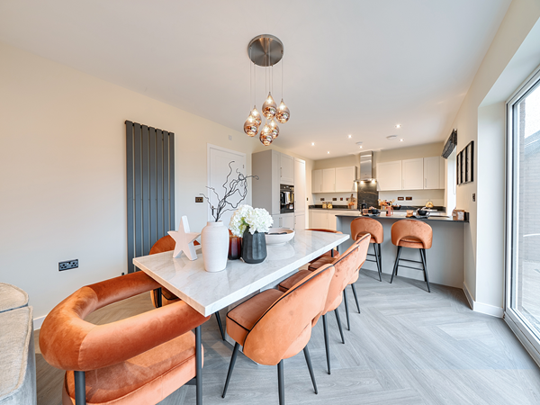 Bright open‑plan dining and kitchen area with a marble dining table surrounded by orange upholstered chairs, decorative pendant lights overhead, and a modern kitchen with white cabinets and a breakfast bar in the background.