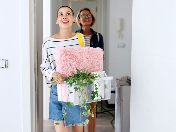 A young woman moving into a home with a box of things, her mother following behind her. 