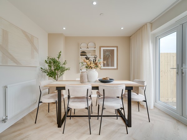 A bright, modern dining room with a light wooden table surrounded by six cushioned chairs in soft beige fabric. The table is decorated with large ceramic vases filled with flowers and greenery. Behind the table is a built‑in shelving nook with neatly arranged bowls and decor pieces, and a framed artwork on the wall. To the right, large glass doors let in natural light and open to an outdoor fenced area. The room features light wood herringbone flooring, neutral walls, and a minimalist, airy aesthetic.