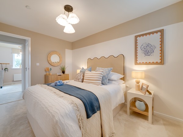 A bright, modern bedroom with a cream bed featuring textured bedding, striped cushions, and a blue throw. The wavy‑edge headboard sits against a two‑tone wall. A bedside table with a lamp, framed photos, and a decorative vase is on the right. A wooden chest of drawers with a round mirror and small décor sits to the left. An open doorway leads to a light‑filled adjoining room.