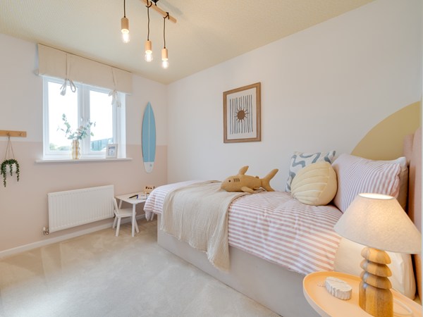 A bright bedroom with a single bed dressed in pink striped bedding, neutral blankets, and decorative cushions. A soft toy sits on the bed. A bedside table with a lamp is on the right, and a small white table with chairs stands near a window on the left. A surfboard‑style wall decoration, framed artwork, and hanging plant add playful touches. The room has light walls, carpeted flooring, and warm pendant lights.