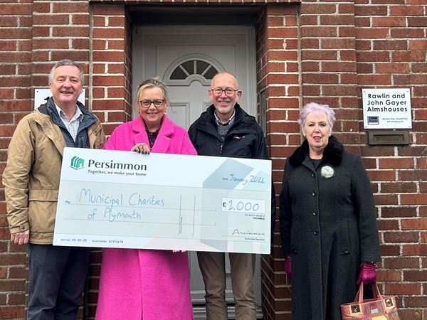 Four people stand in front of a brick building holding an oversized Persimmon cheque for £1,000 made out to “Municipal Charities of Plymouth.” They are positioned in front of a grey door with a sign on the wall reading “Rawlin and John Gayer Almshouses.” One person wears a bright pink coat, another holds a handbag. The group stands close together for the presentation photo.